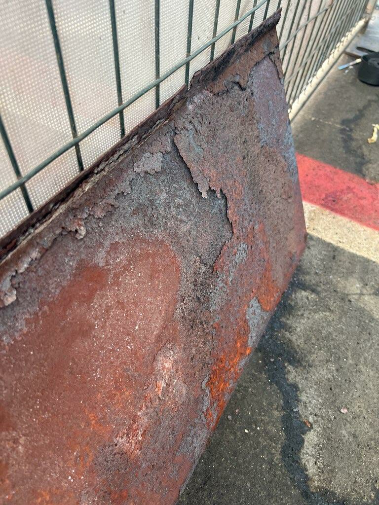 Heavily rusted and flaking metal air deflector panel leaning against a fence